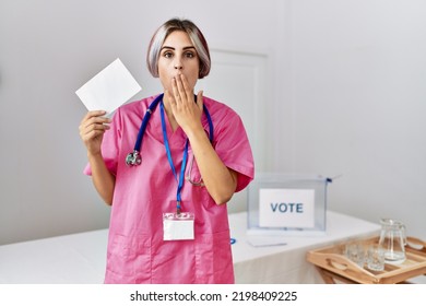 Young Nurse Woman At Political Campaign Election Holding Envelope Covering Mouth With Hand, Shocked And Afraid For Mistake. Surprised Expression 