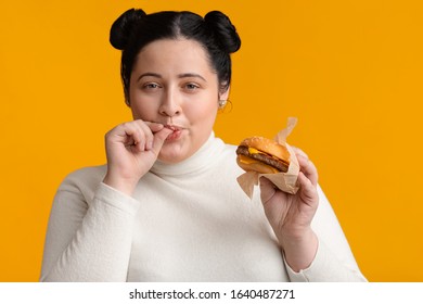 Young Obese Girl Eating Burger And Licking Fingers, Enjoying Fast Food, High Calorie Nutrition