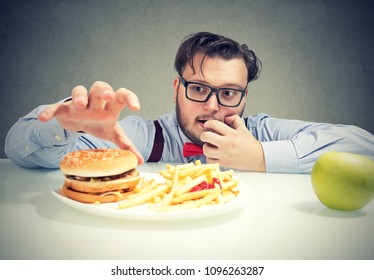 Young obese man in glasses having cravings for cheeseburger with fries instead of healthy green apple. 
