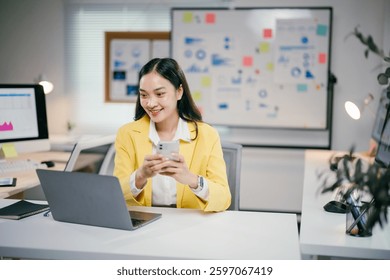 Young office worker using mobile phone and smiling while sitting at desk with laptop in bright workplace