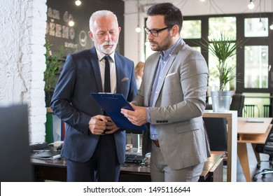 Young And Old Businessmen In Formal Suits Discussing Document On A Meeting In Office