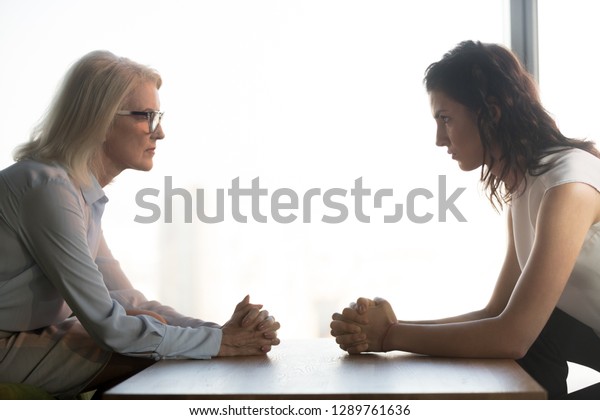 Young and old businesswomen with clasped hands\
looking at each other sitting opposite as rivalry confrontation\
concept, female career success envy and jealousy, generations\
conflict at work concept