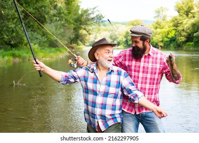 Young And Old Fisherman Standing On The Shore Of Lake With Fishing Rod. Father And Son Enjoy Life. Men Family, Granddad And Drandson Fishing. Mature Man Fisher Celebrate Retirement.