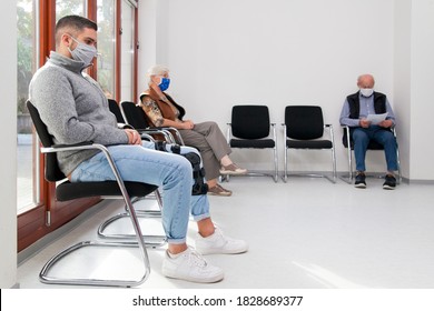 Young And Old People With Face Masks Keeping Social Distance In A Waiting Room Of A Hospital Or Office -  Focus On The Young Man In The Foreground