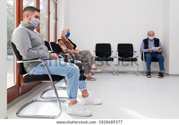 Young and old people with face masks keeping\
social distance in a waiting room of a hospital or office -  focus\
on the young man in the\
foreground