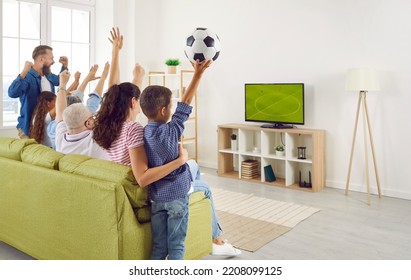 Young And Older Family Members Together Emotionally Cheer For National Team While Watching Football Match At Home. Side View Of Large Family Sitting With Arms Raised On Sofa In Room In Front Of TV.