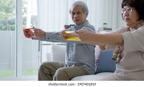 Young older senior asia citizen couple sit on sofa couch with in-home nursing care, assisted living, scrubs nurse use resistance band exercise for senior patient in physiotherapy treatment at home.