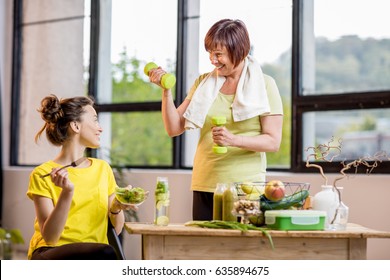 Young And Older Women Exercising With Dumbbells And Eating Healthy Food Indoors On The Window Background