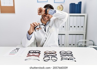 Young Optician Man Holding Optometry Glasses Smiling Cheerful Playing Peek A Boo With Hands Showing Face. Surprised And Exited 