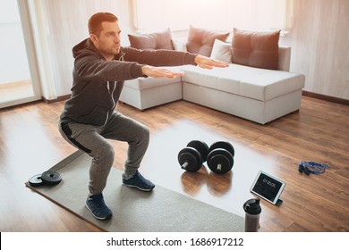 Young Ordinary Man Go In For Sport At Home. Real Picture Of Egular Guy Doing Squats With Stretch Hands Forward. Beginner Or Amateur Has Workout In Apartment. Sport Equipment On Floor