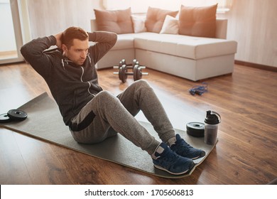 Young ordinary man go in for sport at home. Hardworking freshman sit on mat and do abs excercise. Not easy to start workout alone in apatment. Beginner in action. Sport equipment on floor. - Shutterstock ID 1705606813