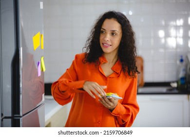 Young Organized Female With Curly Hair In Casual Clothing Making To-do And Checklist On Sticky Notes As Reminder And Placing Them On Refrigerator In Kitchen At Home