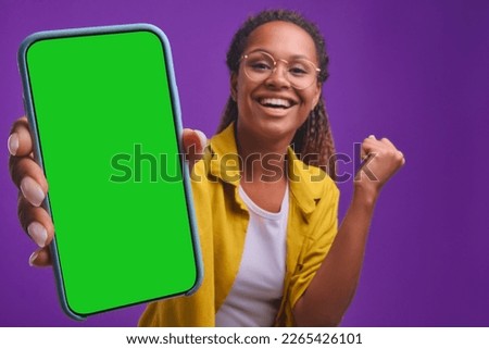 Young overjoyed African American woman stretches out hand with smartphone showing off record in mobile game or result in application with intelligence test stands in purple studio. phone green screen