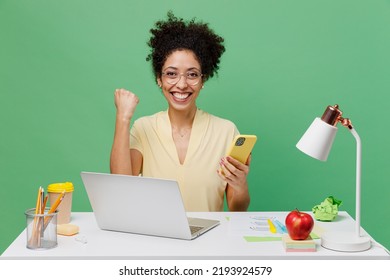 Young Overjoyed Employee Business Woman Of African American Ethnicity In Shirt Sit Work At White Office Desk Pc Laptop Hold Use Mobile Cell Phone Do Winner Gesture Isolated On Plain Green Background