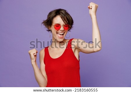 Young overjoyed happy woman 20s she wear red tank shirt eyeglasses doing winner gesture celebrate clenching fists say yes isolated on plain purple background studio portrait. People lifestyle concept