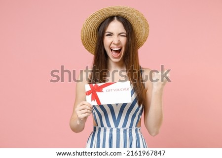 Young overjoyed happy woman in summer clothes striped dress straw hat hold in hand gift voucher flyer mock up do winner gesture clench fist isolated on pastel pink color background studio portrait.
