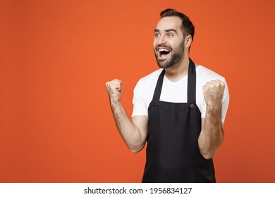 Young Overjoyed Man Barista Bartender Barman Employee In Black Apron White T-shirt Work In Coffee Shop Do Winner Gesture Clench Fist Isolated On Orange Background Studio Small Business Startup Concept