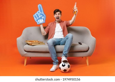 Young overjoyed man football fan in shirt support team with soccer ball sit on home sofa watch tv live stream hold pizza foam glove finger drink beer isolated on orange background People sport concept