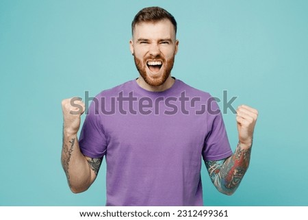 Young overjoyed man he wears purple t-shirt doing winner gesture celebrate clenching fists say yes isolated on plain pastel light blue cyan background studio portrait. Tattoo translates life is fight