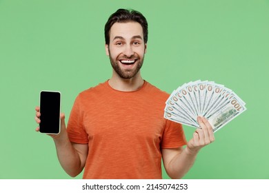 Young overjoyed man in orange t-shirt holding fan of cash money in dollar banknotes mobile cell phone with blank screen workspace area isolated on plain pastel light green background studio portrait.
