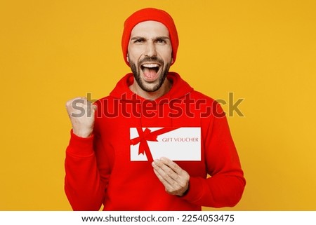 Young overjoyed surprised happy fun man wears red hoody hat look camera hold gift certificate coupon voucher card for store do winner gesture isolated on plain yellow color background studio portrait