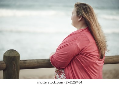 Young overweight caucasian adventurous teenage girl with blonde hair looking out at the ocean, while leaning on the wooden railing along the beach