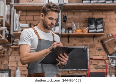 Young owner in an apron writing on clipboard while checking products in restaurant. Man looking on checklist and doing inventory. Small business concept. Entrepreneur in hospitality industry. - Powered by Shutterstock