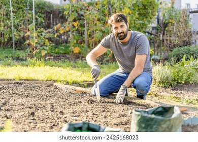 Young pakistani eastern happy smiling bearded farmer in jeans, t-shirt and protective gloves digging up the ground to harvest potatoes or plant vegetable seeds in horticultural garden on sunny day off