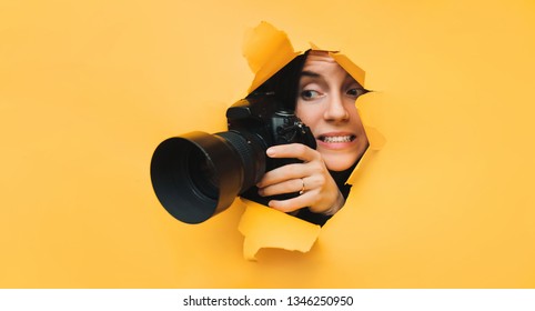 A young paparazzi girl holds a reflex camera and looks through a torn hole in yellow paper. The concept of embarrassment, shame for what he saw. Copy space.