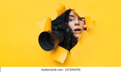 A young paparazzi girl with a reflex camera looks out from cover and looks at what is happening with her mouth open. Yellow paper, torn hole. Tabloid press. In search of the plot for photo stocks.