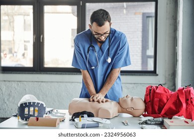 young paramedic in blue uniform and eyeglasses practicing chest compressions on CPR manikin near defibrillator and first aid kit during medical seminar, life-saving skills development concept