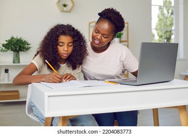 Young Parent Helping Child With School Homework. Happy Beautiful African American Mother And Her Little Daughter Sitting At Desk At Home, Using Modern Laptop Computer And Writing In Notebook