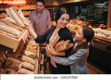 Young parents and daughter in grocery store. Cheerful mother and girl pick upb bread and rolls together. They smile to each other. Man stand behind with trolley. - Powered by Shutterstock