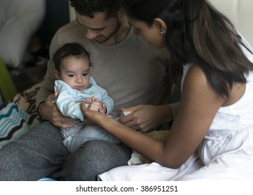 Young Parents Giving Lots Of Attention To Their Baby. They Are All Sitting On A Bed At Home.