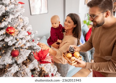 Young parents having fun decorating Christmas tree with their cute little baby girl, placing ornaments and decorating home for winter holiday season