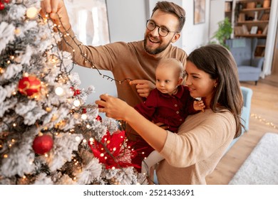 Young parents having fun decorating Christmas tree with their cute little baby girl, placing Christmas lights on it while decorating home for winter holiday season - Powered by Shutterstock