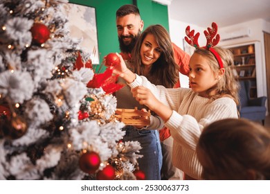 Young parents having fun decorating Christmas tree with their daughters, placing Christmas lights and ornaments on it while decorating home for winter holiday season - Powered by Shutterstock