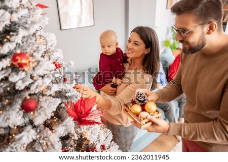 Young parents having fun decorating Christmas tree with their cute little baby girl, placing ornaments and decorating home for winter holiday season