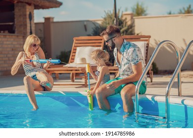 Young parents having fun playing with their daughter by the swimming pool splashing water on each other, enjoying hot sunny summer day outdoors and relaxing while on a vacation