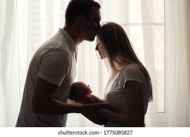 Young parents are holding their newborn baby in their arms. The husband kisses his wife. The concept of a happy young family.