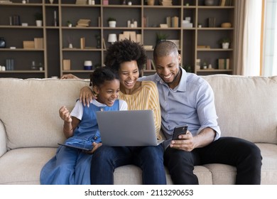 Young parents and little 7s daughter sit on sofa with diverse devices watching on cellphone funny videos laughing looking overjoyed. African family enjoy modern tech, online humorous content concept - Powered by Shutterstock