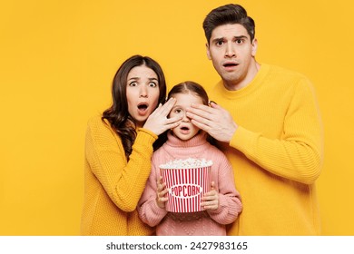 Young parents mom dad with child girl 7-8 years old wear pink casual clothes watch movie film eat popcorn cover kid eyes because of bad scene isolated on plain yellow background. Family day concept