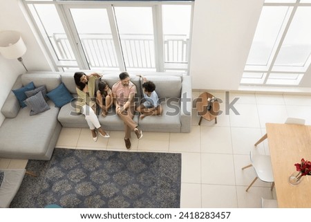 Young parents and two preteen sibling children sitting on couch in spacious apartment, talking, enjoying family home leisure in living room with sofa, carpet, dining table