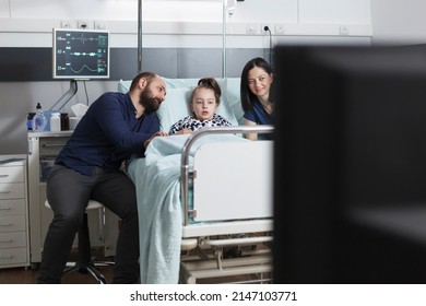 Young parents and unwell daughter watching television channel while sitting in pediatric clinic ward consultation room. Family spending time together in clinic patient room while enjoying tv show.
