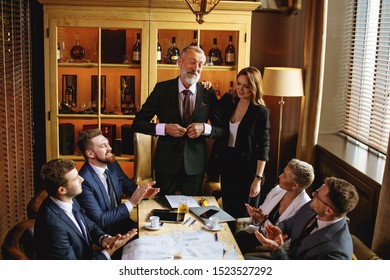 Young Partners Happily Applauding And Looking At His Old Mentor, Businessman Buttoning Black Blazer, Beautiful Colleague Standing With A Smile By Side, Not Looking To Camera