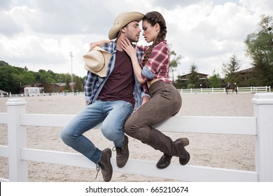 young passionate cowboy style couple kissing while sitting on fence at ranch