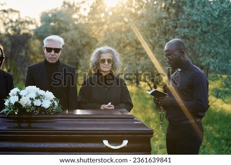 Young pastor reading verses from Holy Bible while standing in front of coffin with closed lid and mourning relatives of passed away person