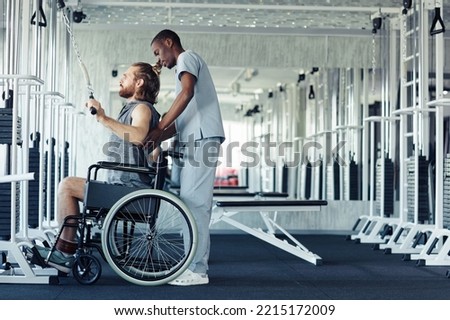 Young patient with disability sitting in wheelchair and training his back on equipment with the help of doctor