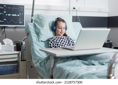 Young Patient Girl Sitting In Pediatric Hospital Bed While Watching Funny Cartoons. Sick Little Child Using Computer To Watch Comical Content On Internet While Sitting In Pediatric Clinic Ward Room.