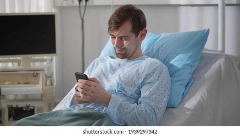 Young Patient Lying In Bed And Using Smartphone While Recovering In Hospital. Portrait Of Sick Man Resting In Bed And Surfing Internet On Mobile Phone In Hospital Ward